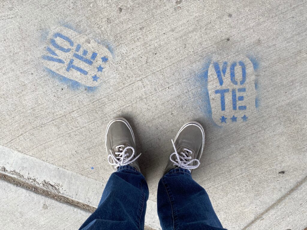 The legs and feet of a person in blue jeans and canvas tennis shoes stands looking down at two blue paint stenciled "Vote" on concrete.