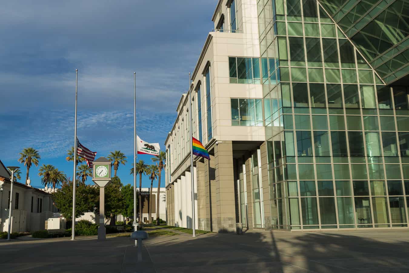A United States flag, a state flag of California, and a Pride flag are blowing in the wind outside a city building in Fairfield, CA.