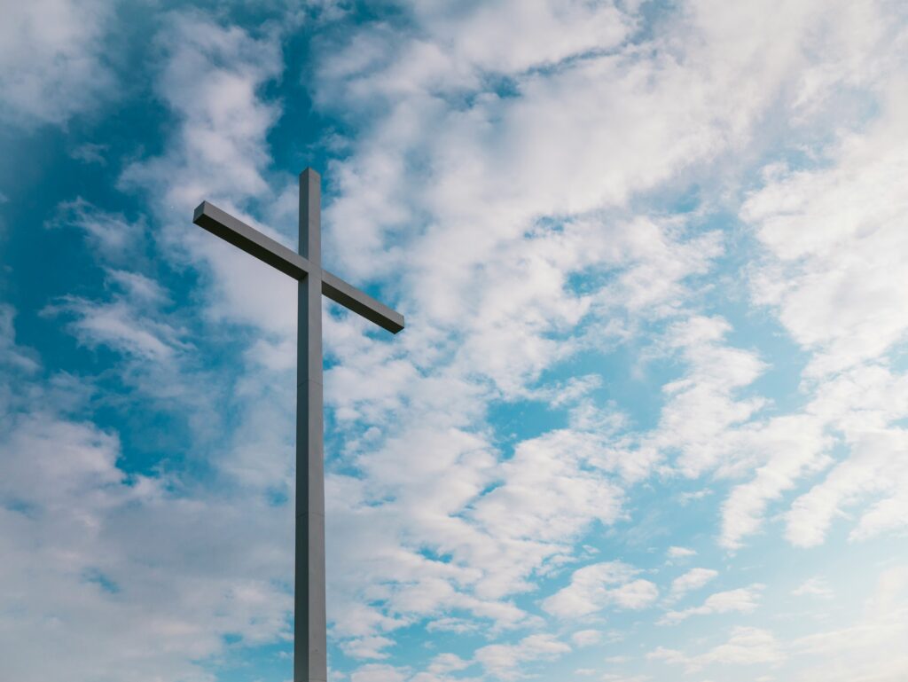 A thin wooden cross is show with a blue sky with mottled white clouds.