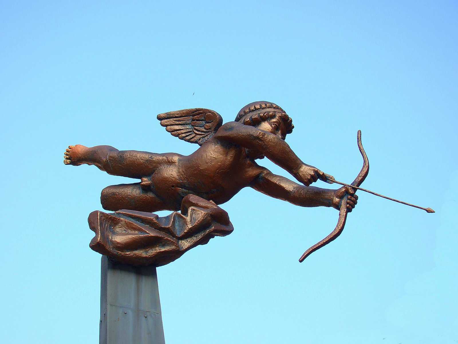 A bronze casting of Cupid shooting an arrow from on a spire.