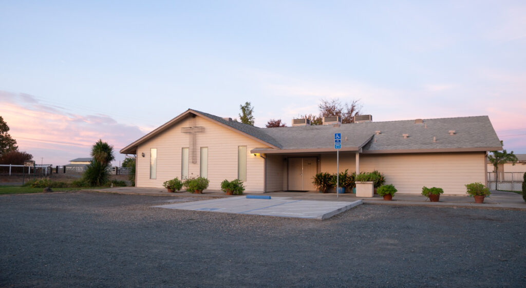 When the Church Forgets Scripture This image features a church building with a cross on the front, set in a gravel parking lot with potted plants and a serene sunset sky.