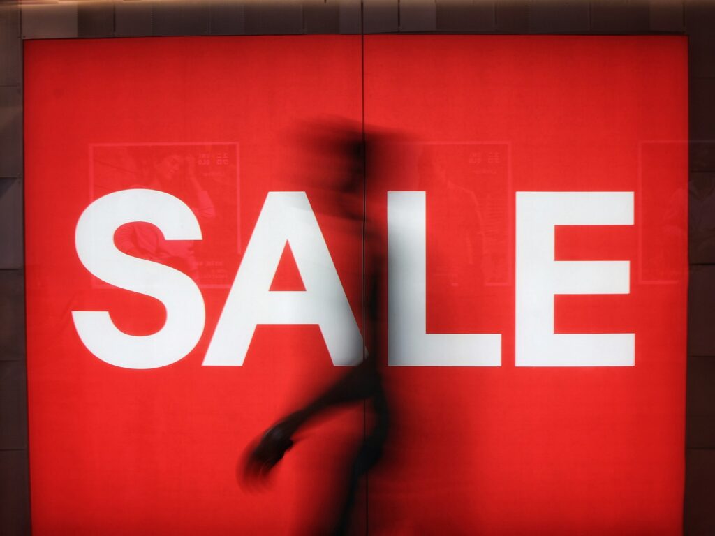 Behind Life Surge: Faith, finances, and a founder’s controversial past A vibrant red sale sign in a retail environment with a blurred silhouette of a shopper walking behind.