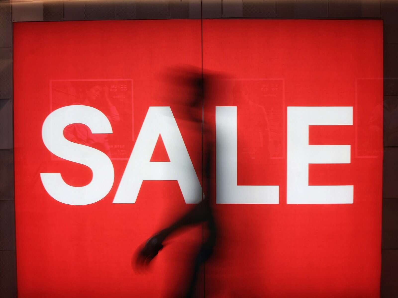 A vibrant red sale sign in a retail environment with a blurred silhouette of a shopper walking behind.