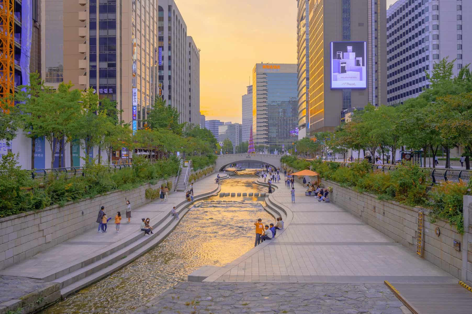 This image depicts a bustling urban river corridor lined with trees and modern high-rise buildings, at sunset in Seoul, South Korea