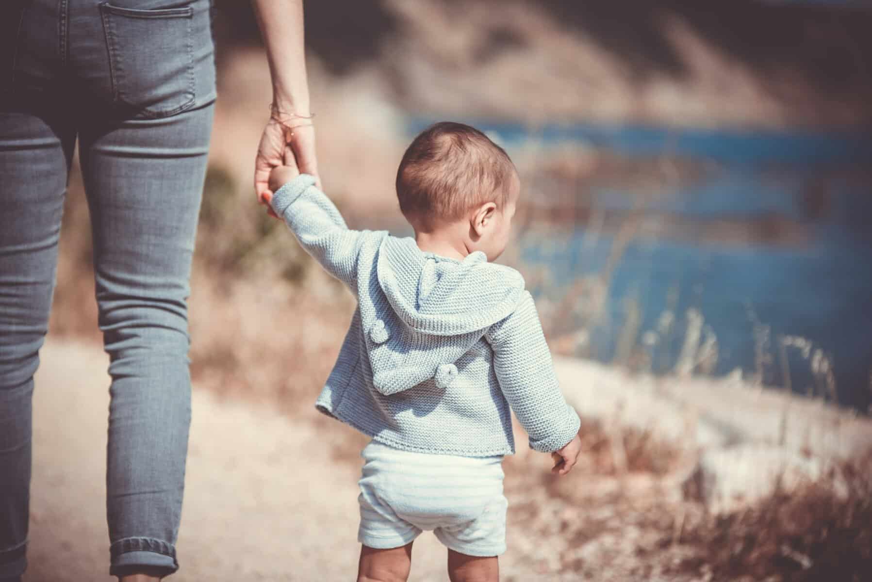 A woman holding a toddler's hand by a lake.