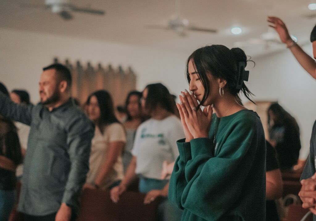 A woman with hands in prayer, eyes closed, participating in a faith-based worship service.