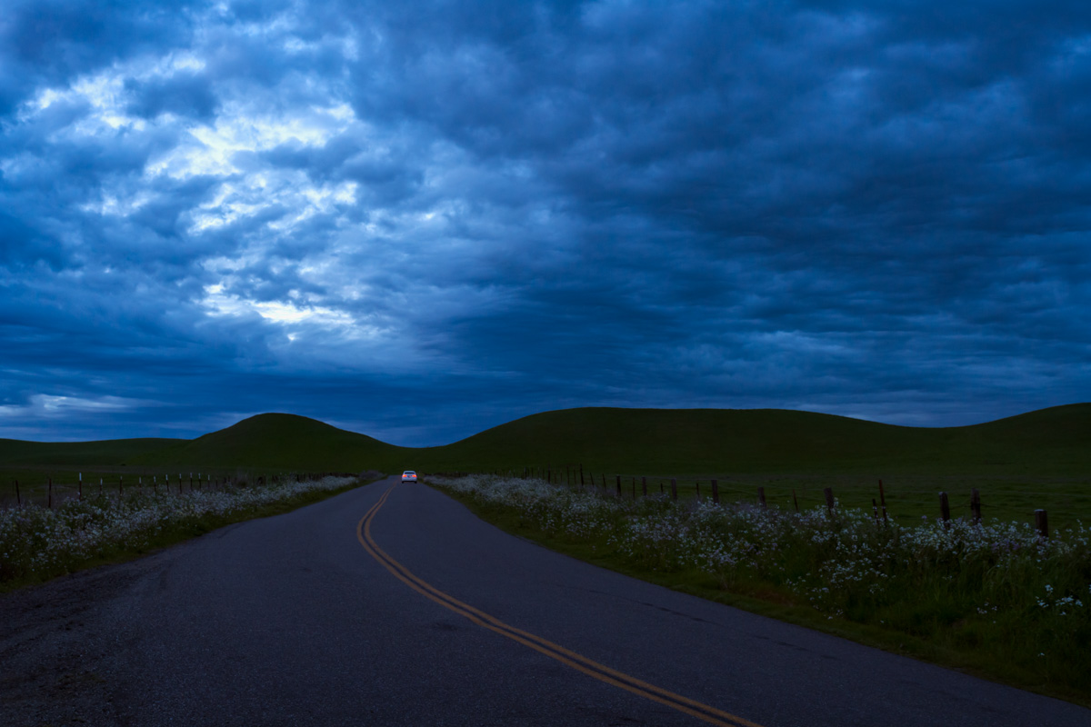 A lone car driving at dusk toward hills on a stormy evening.