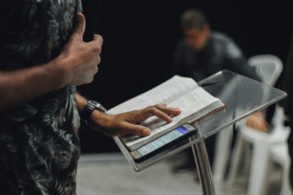A man standing at a lectern with an open Bible and holding microphone.