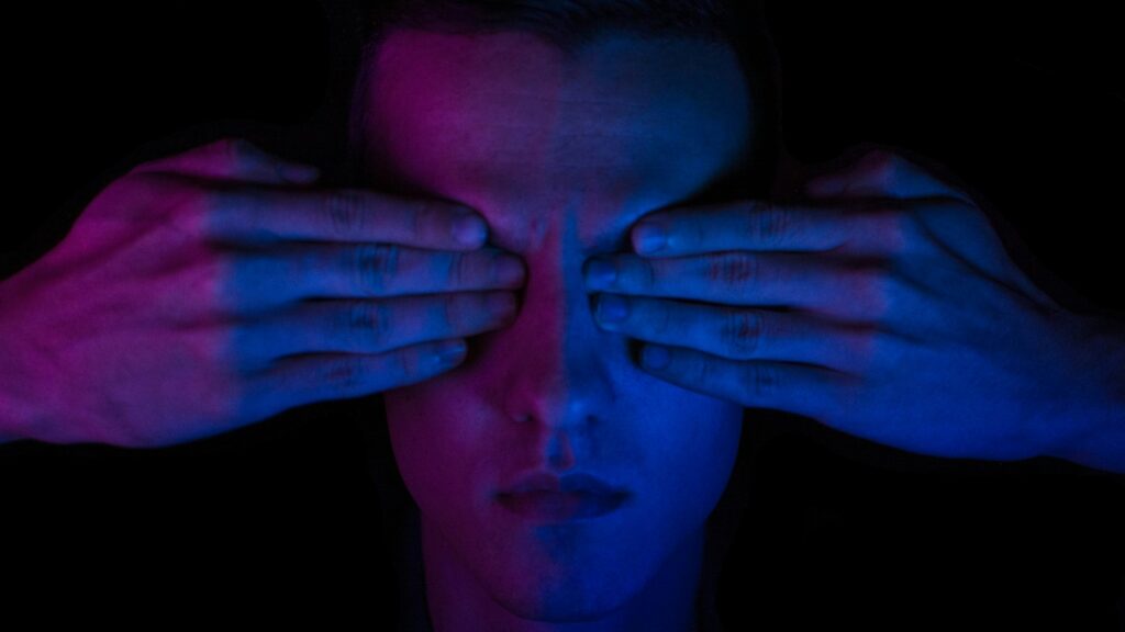 A person in low blue and red light with their eyes covered by hands.