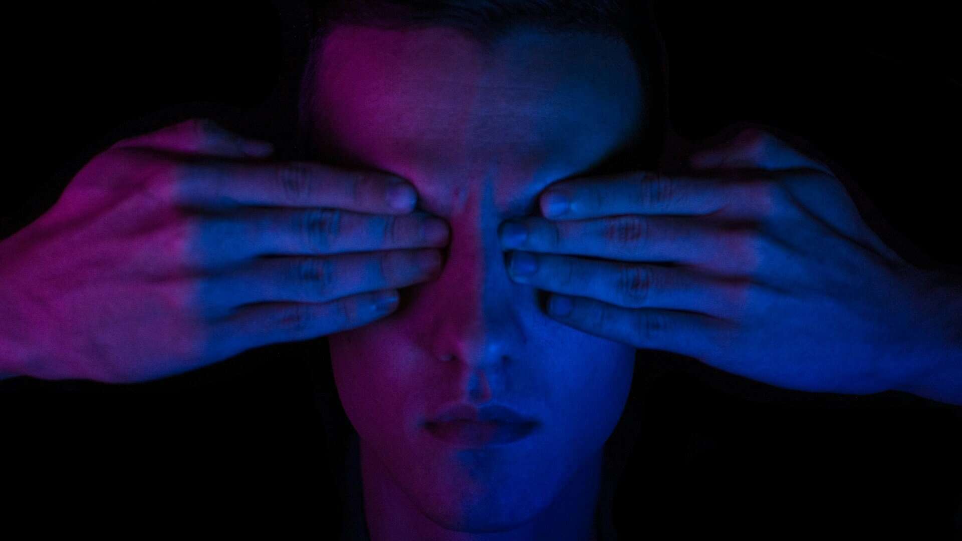 A person in low blue and red light with their eyes covered by hands.