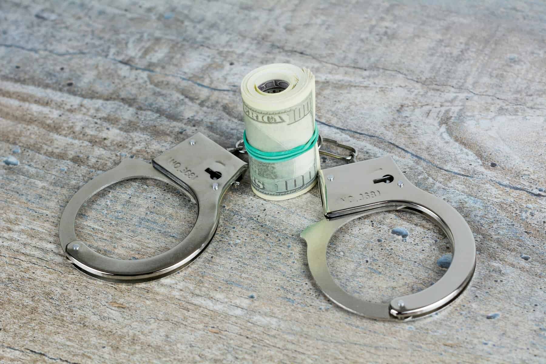 Handcuffs are shown with a large roll of U.S. 100 dollar bills.