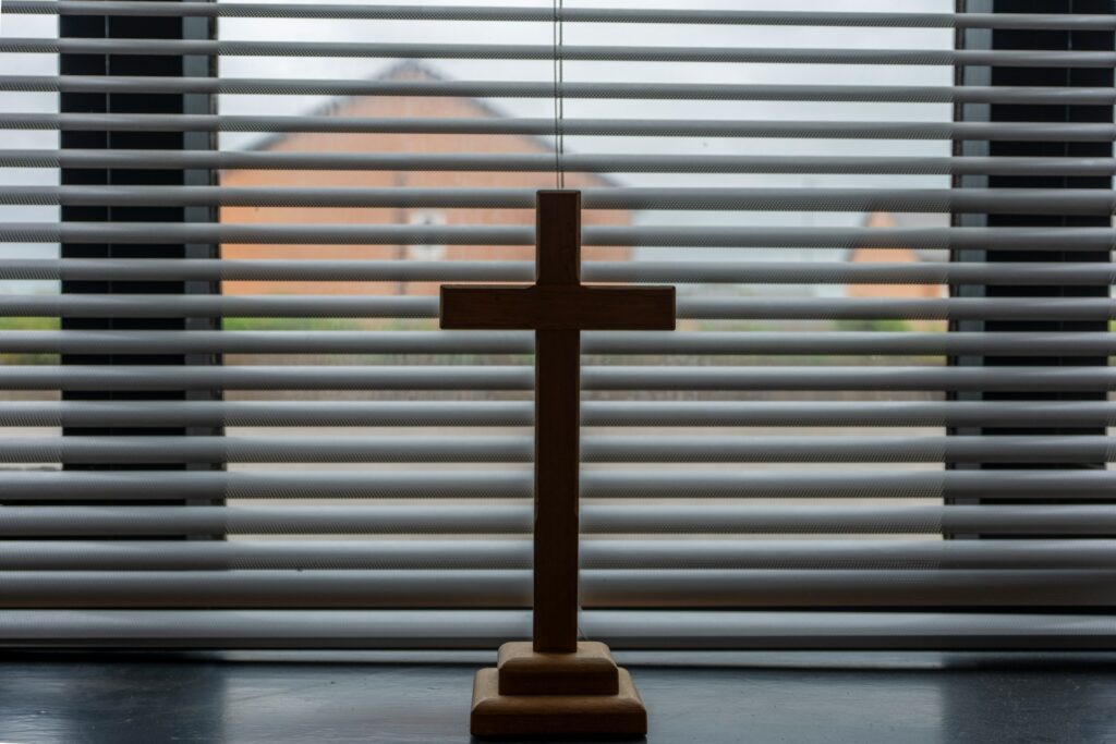 A wooden cross in a window sill in front of miniblinds.