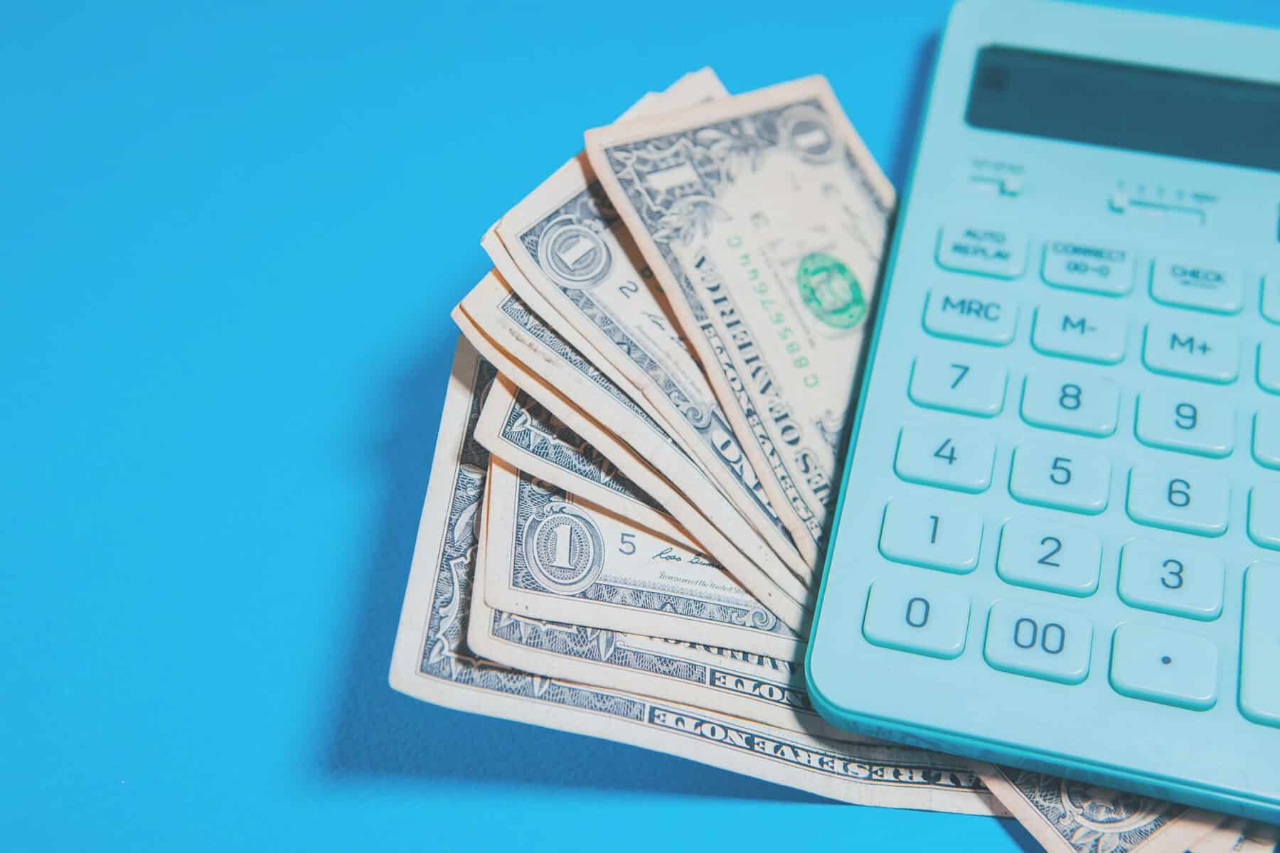 A fanned stack of dollar bills and a blue calculator sit on a soft blue background.