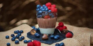 A glass dessert cup sits on a square of burlap and is filled with vanilla and chocolate pudding and topped with blueberries and raspberries.