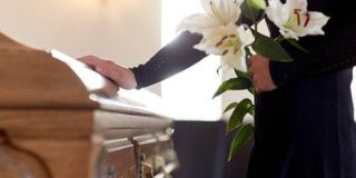 A woman dressed in black is shown with a hand on a casket and holding a bouquet of white flowers.