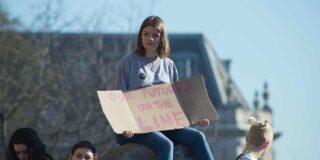 A teen girl sits on a boulder holding a cardboard sign that says in red "our future's on the line".