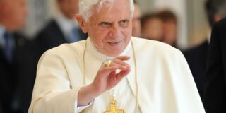 Pope Emeritus Benedict XVI is shown in off white Pope robes and gesturing to a crowd.