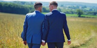 Two men dressed in blue suits hold hands and walk away from camera in a pastoral setting.