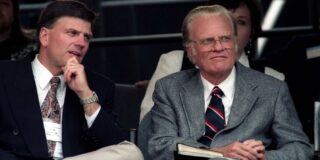Billy Graham is shown seated in an auditorium type setting and wearing a grey suit.