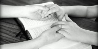 Black and white image shows two people's hands a forearms. The people are holding hands resting on a bible that is open on a table top.