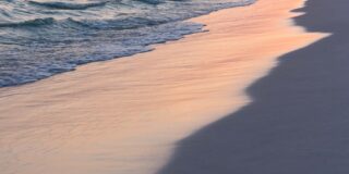 Ocean waves are shown sliding back into off of sand with the newly soaked sand reflecting the pink sunset light.