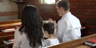 A mother, child, and father are seen in a pew from the back and in that order from a church aisle.