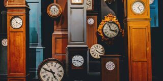 Grandfather clocks and mantle clocks of various colors and sizes are arranged all together in a darker room.
