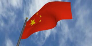 A red Chinese flag on a pole is seen from below with a blue sky and thin white clouds above.