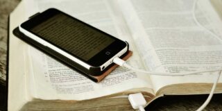 A smart phone is seen sitting on a bible with its cord appearing to be plugged into the open bible.