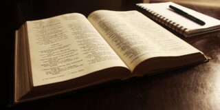 A Bible is shown open faced in soft light with a notepad and with a pen next to it.
