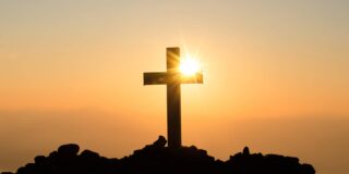 A thick beam cross is shown on a hill with the sun setting behind it.