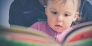 A toddler sits on an adults lap as they look at an open book that is out of focus and between the camera and readers.