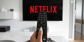 A TV with the Netflix logo hangs on a white wall with a woman's manicured hand holding a remote between the camera and the tv.