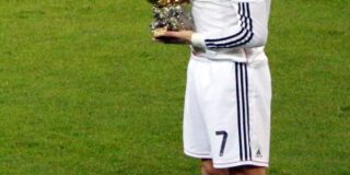 Soccer player Cristiano Ronaldo is seen on the field in a white uniform and holding a ball trophy.