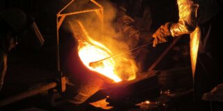 A metal worker wearing protective heavy clothing holds an iron in molten metal.