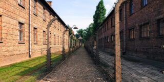 A barbed wire fence and long brick buildings are shown at Auschwitz.