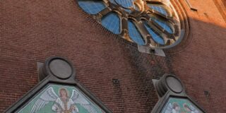 A historic red brick church facade in Amsterdam with tile work and a rose window are seen close up from below with camear angled up.
