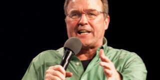 Mike Bickle is seen in a green shirt and speaking into a microphone he's holding.