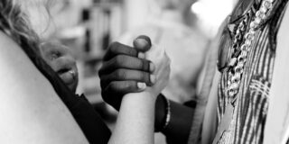 A white person and a Black person are shown facing each other and clasping hands at chest level.